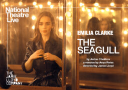 Actress Emilia Clarke stands in front of a brightly lit mirror, with the title of the play 'The Seagull' in white text