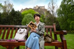 Actress Sally Hawkins sits on a wooden bench reading a book in front of a castle