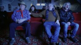 A group of old men and women sit in cinema chairs watching a film