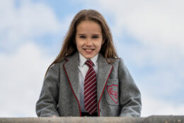 A young girl in school uniform looks directly towards the camera against a blue sky background